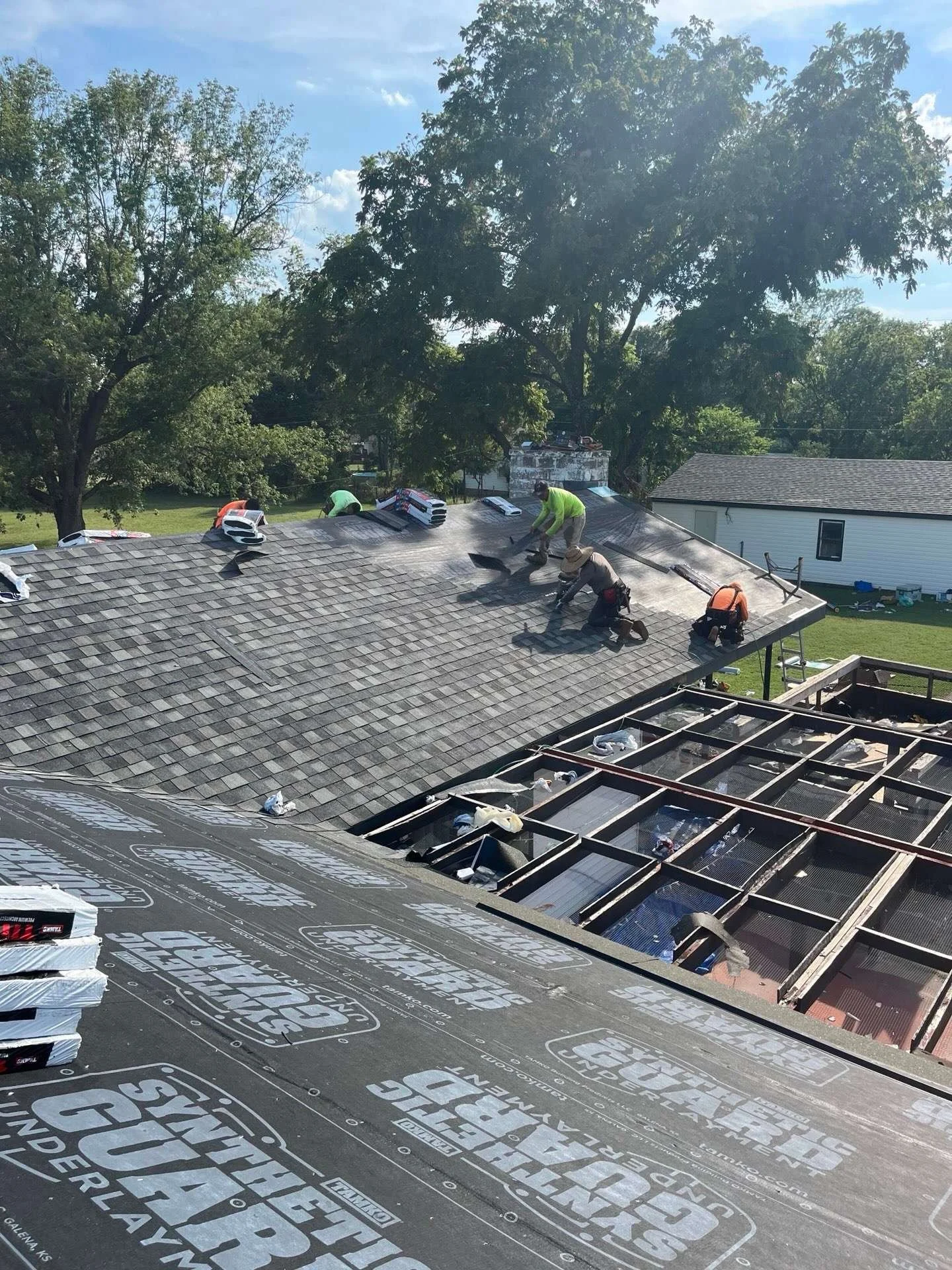 Workers installing new roofing on a house with gray shingles. Some workers are on the roof, while others are where the roof was removed. The house is in a residential area with trees and a blue sky.