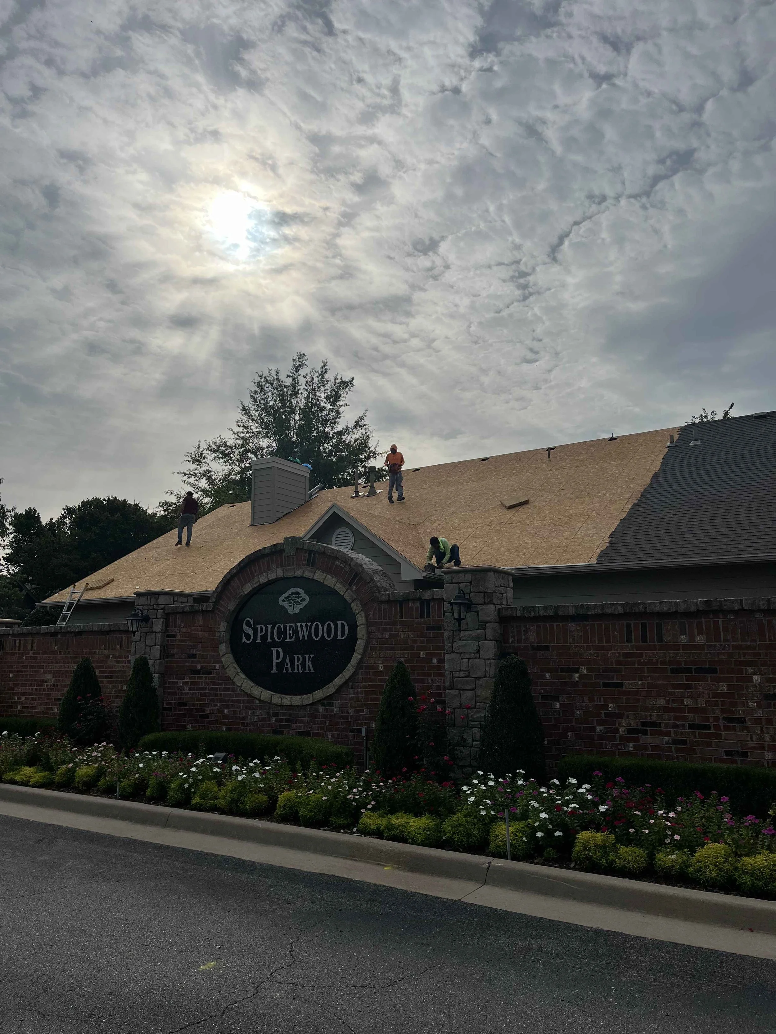 The entrance sign of Spicewood Park with a brick wall, flowers in front, and a roof with workers repairing it under a cloudy sky with the sun peeking through.