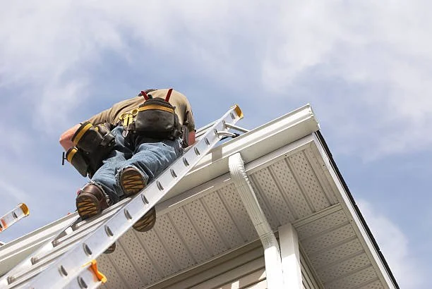 A person working on a ladder on the exterior of a house, with a cloudy sky in the background.