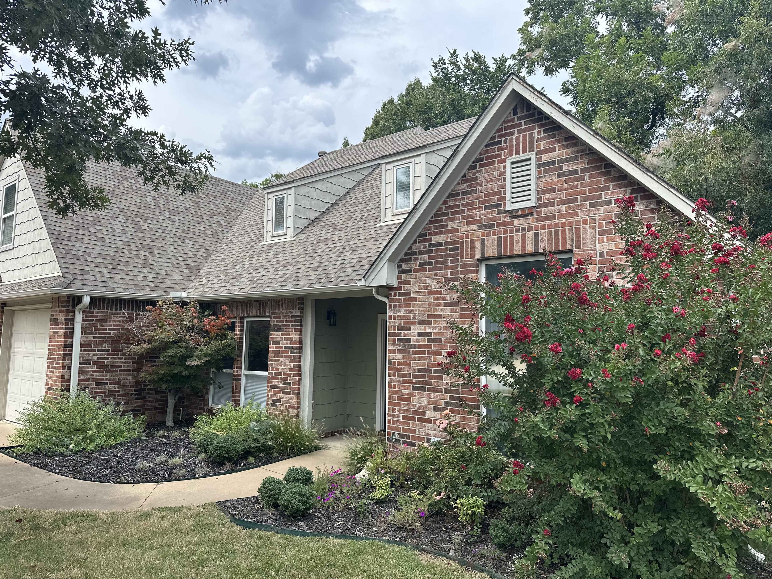 A two-story house with brick and gray siding exterior, with a sloped roof, surrounded by green trees and a landscaped yard with bushes and flowers.