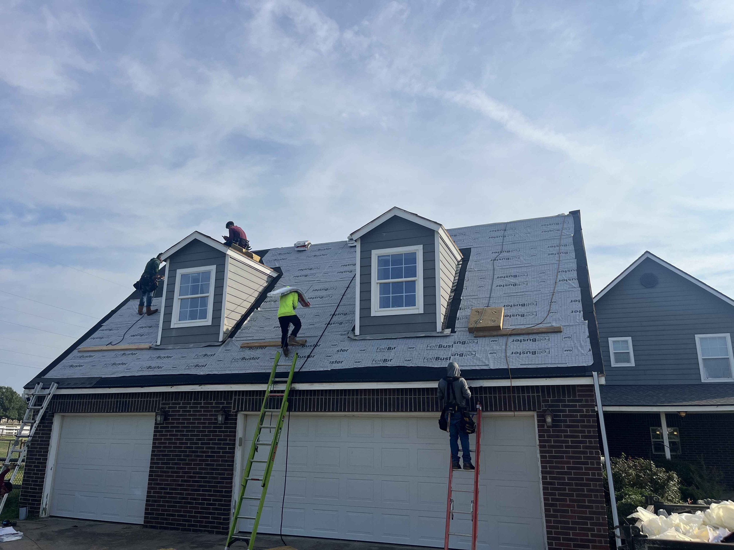 Workers installing roofing on a house, with some on the roof and others on ladders or scaffolding, using various tools.