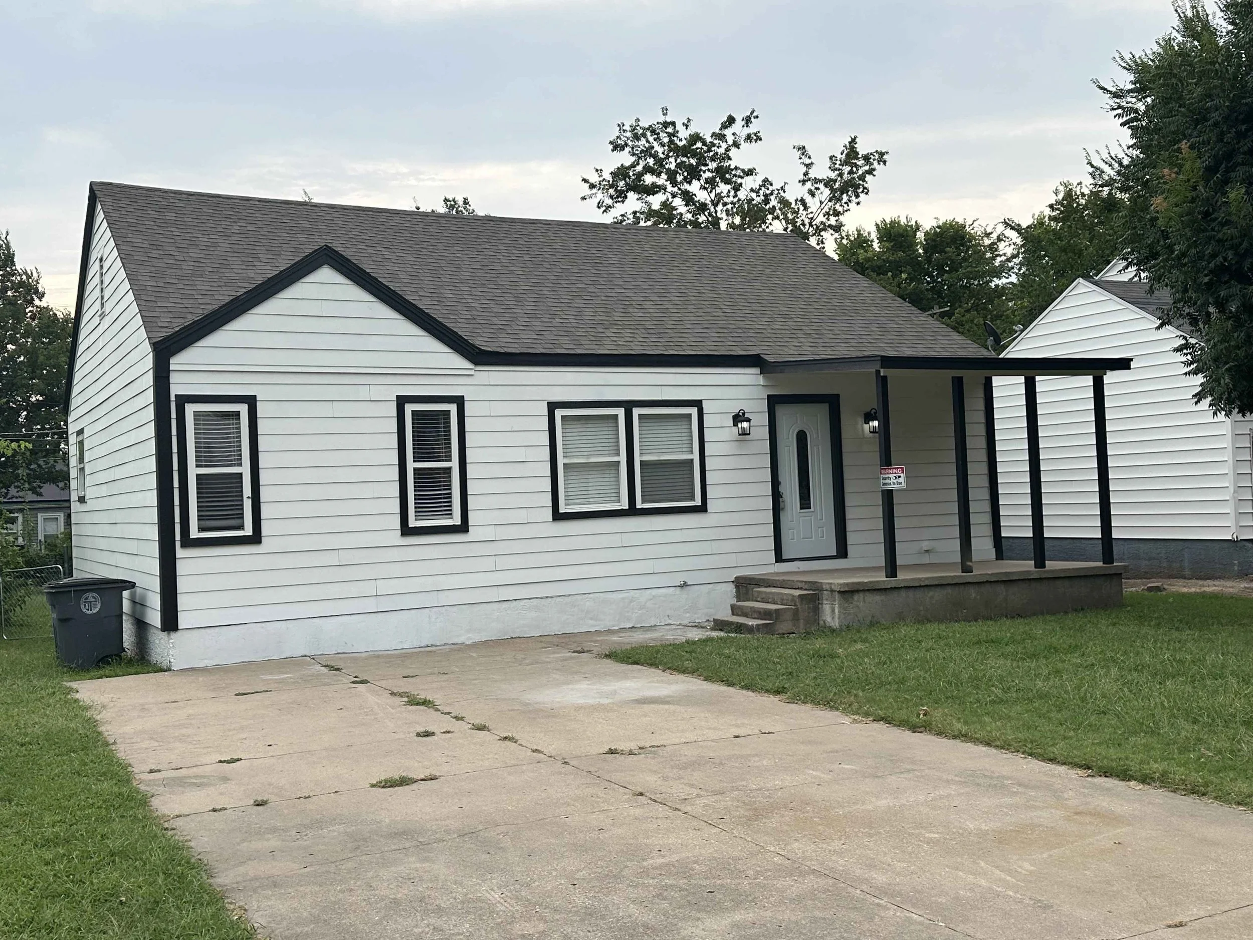 A white house with black trim, a gray roof, and a small porch with stairs. There is a lawn, a sidewalk, a trash bin, and trees in the background.