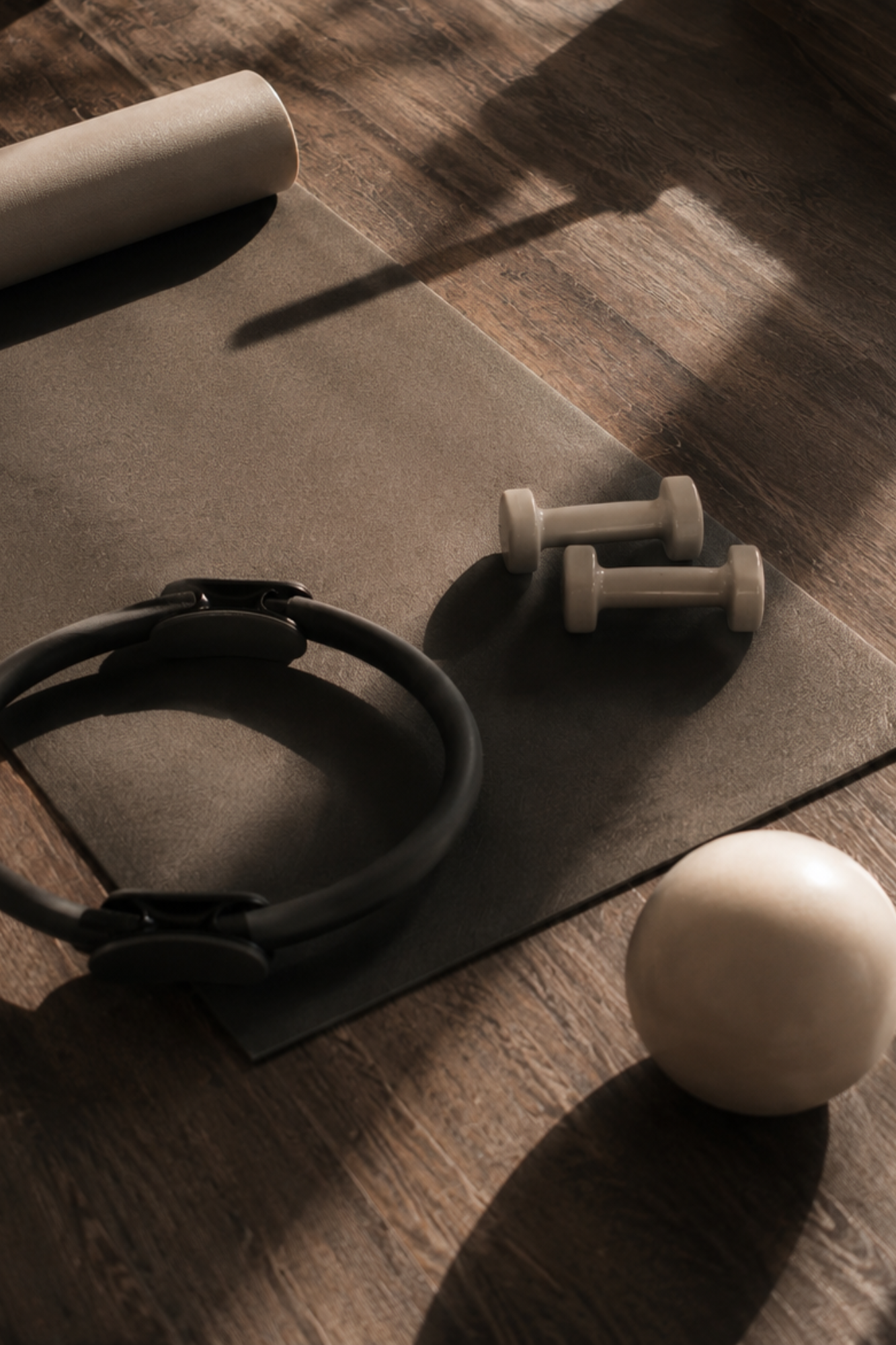 A yoga mat, a pair of dumbbells, a kettlebell, and a medicine ball on a wooden floor with sunlight and shadows.