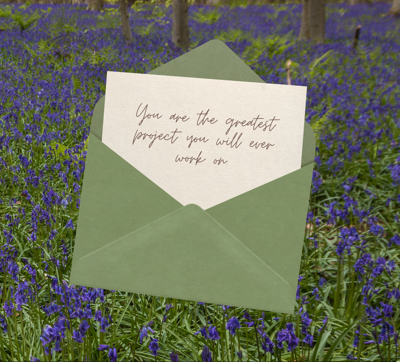 A green envelope holding a note that says, "You are the greatest project you will ever work on," in a field of purple flowers and trees in the background.