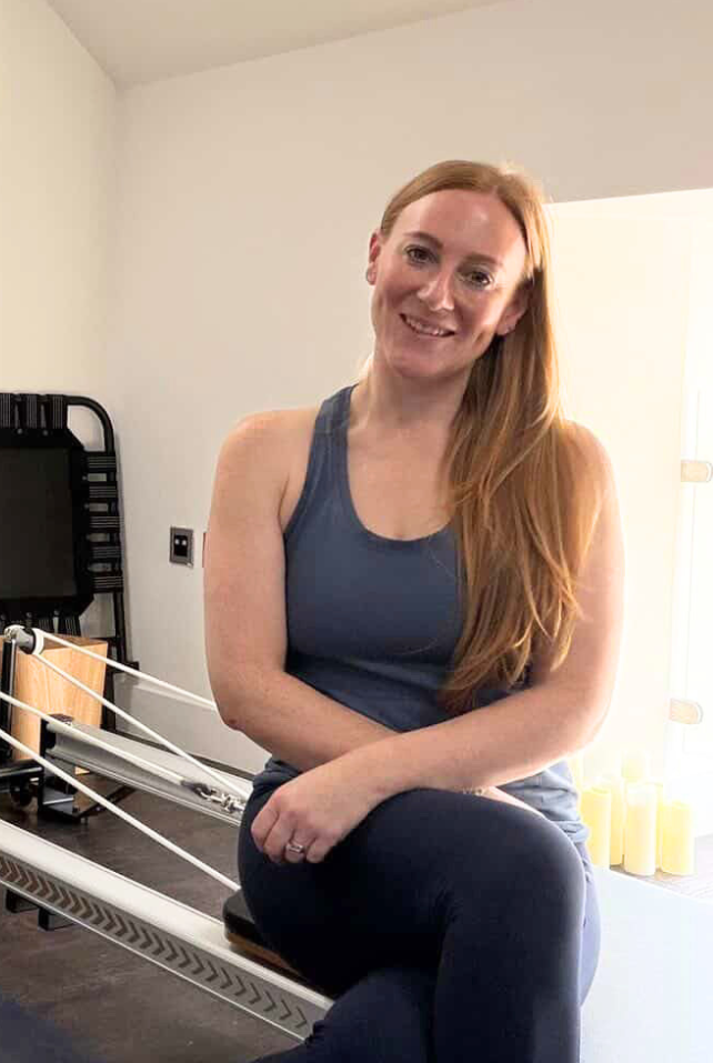 Instructor, Stephanie Proudfoot, sitting on a Pilates reformer machine in a bright room.