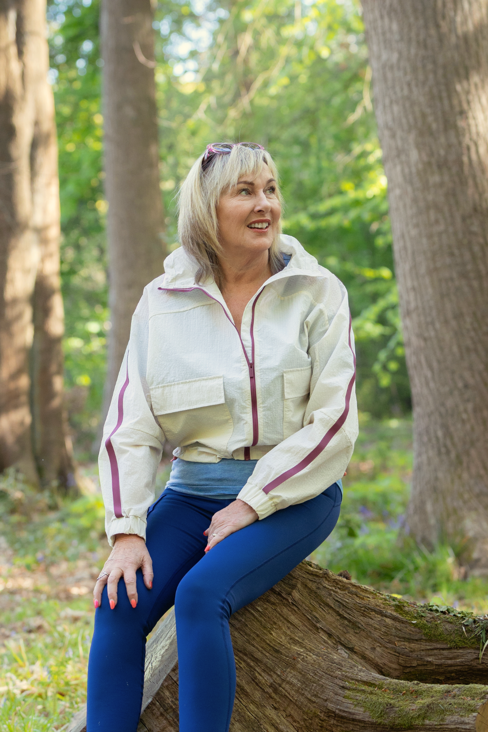 Founder of The Wellness Space, Lorraine Young, sitting on a fallen log in a forest, smiling and looking to her right, wearing a white windbreaker, blue pants, and sunglasses on her head.