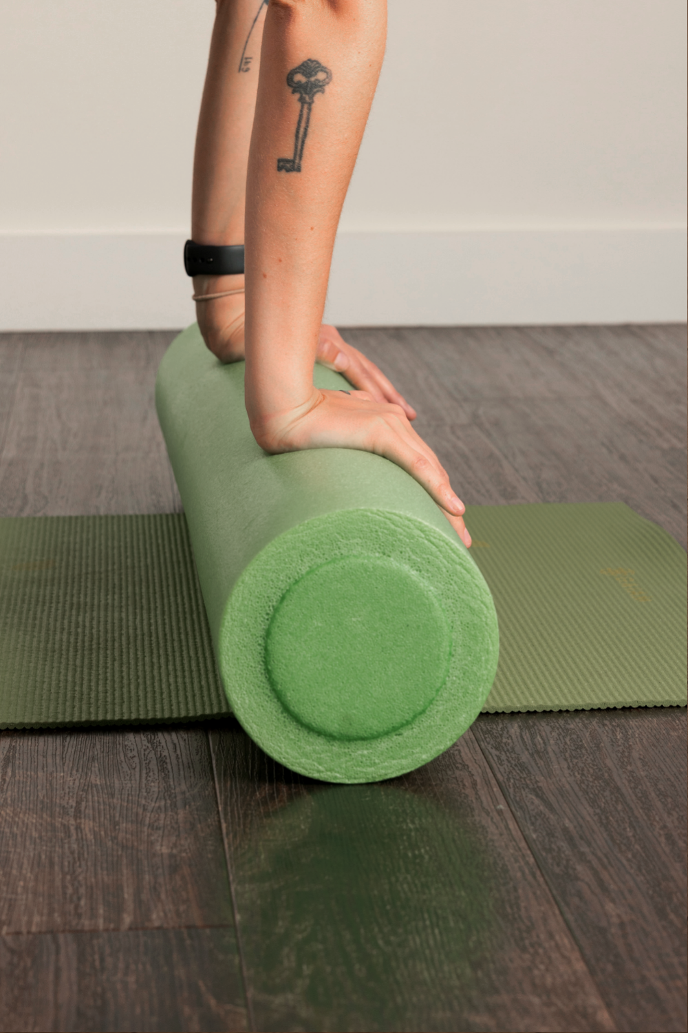 Person performing pilates on a green foam roller, with hands on the roller, on a green yoga mat indoors.