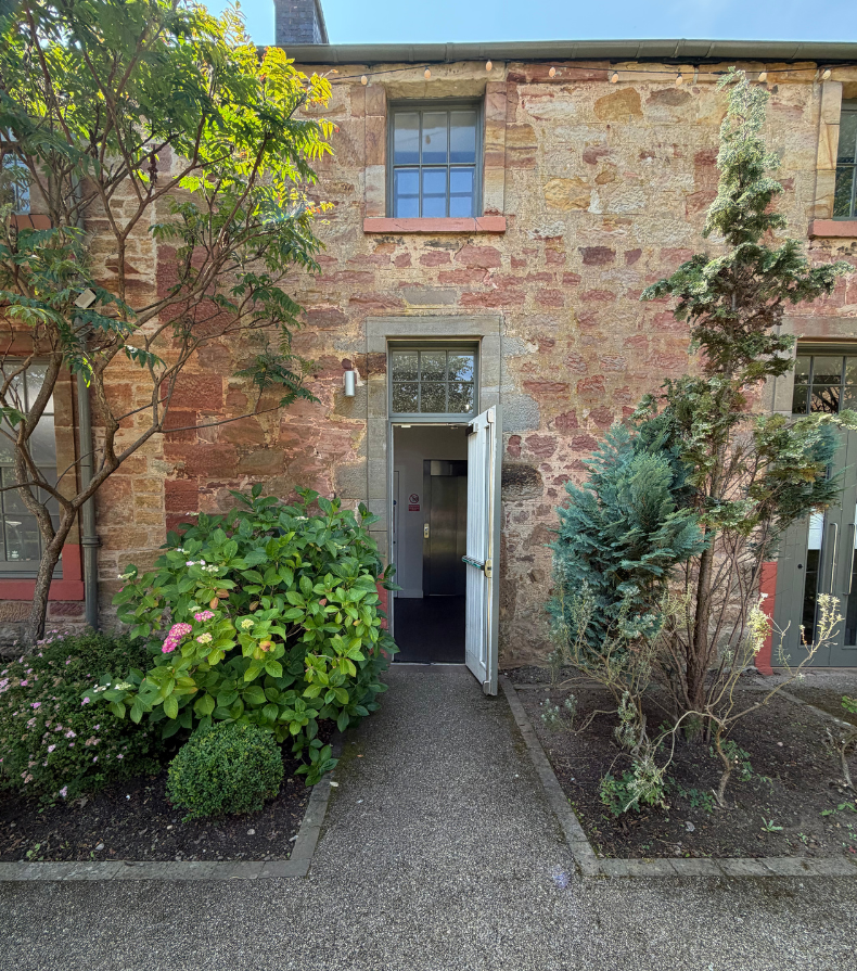 The Wellness Space studio entrance, a brick building with a door slightly open, surrounded by green bushes and trees, with a window above the door.