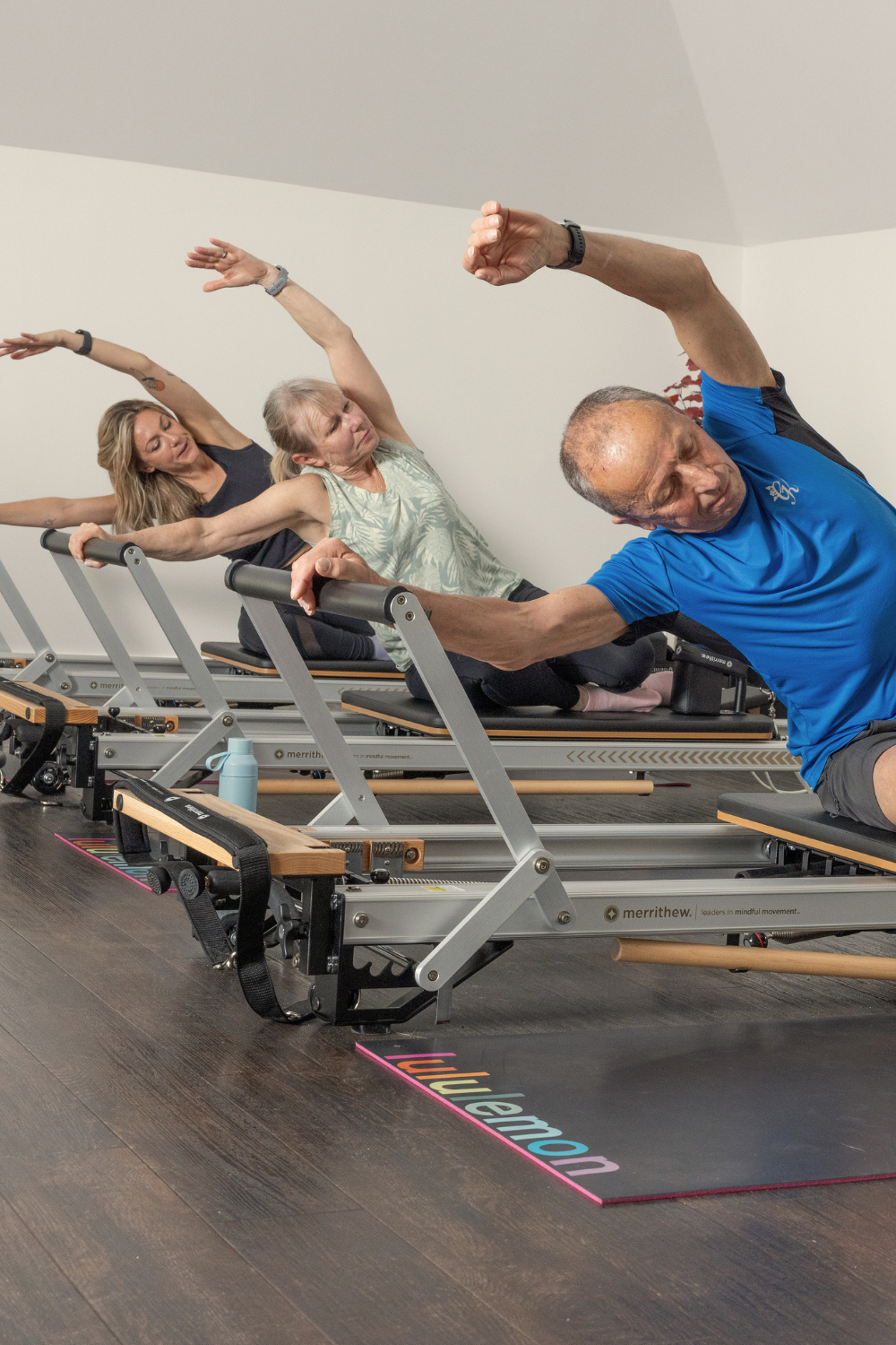 Two women and one man participating in a Pilates class on reformer machines, stretching their upper bodies with their arms raised and leaning to the side in a room with white walls and wooden flooring.
