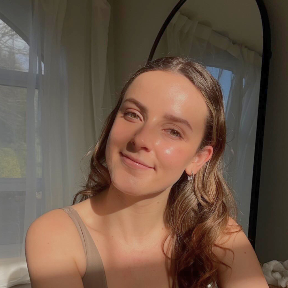 Instructor, Sophie Newman, with long wavy brown hair smiling at the camera, inside a room with sheer curtains and a window in the background.
