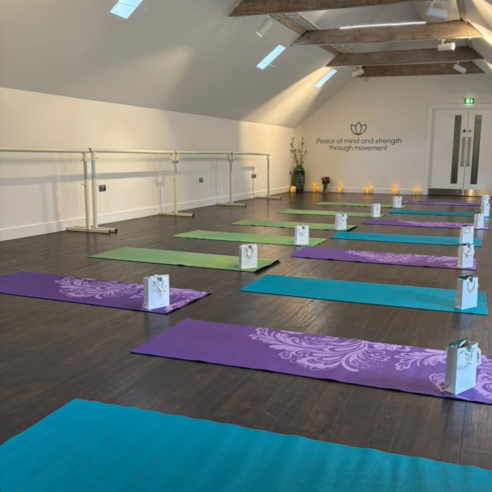 Yoga mats in The Wellness Space studio in purple, teal, and green colors are neatly arranged on a wooden floor in front of a ballet barre. Each mat has a small gift bag and a face mask placed at the top.