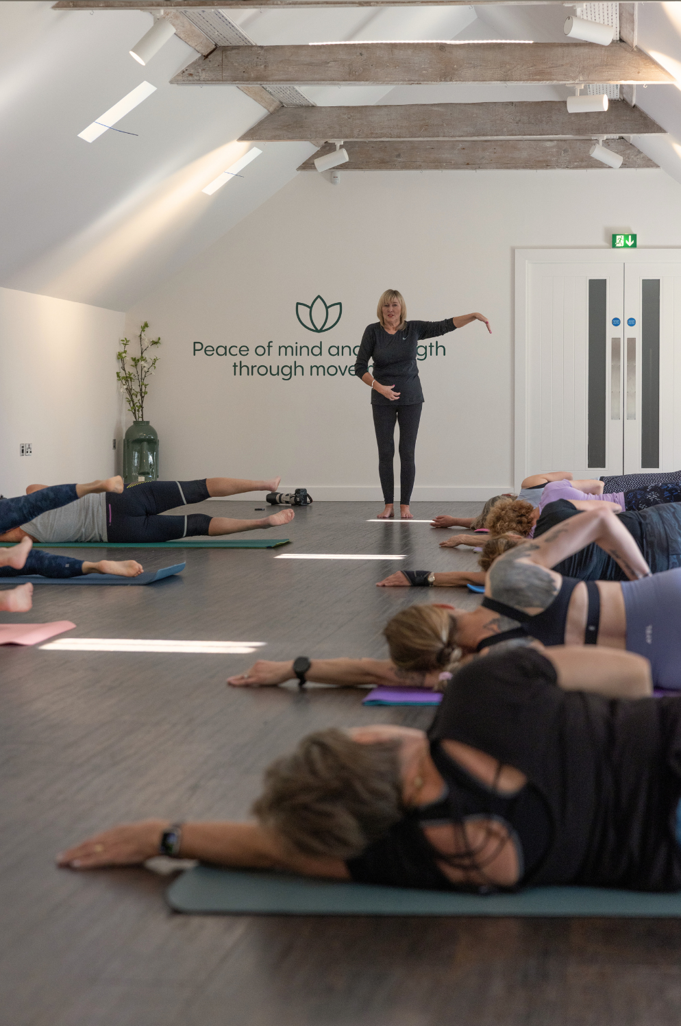 Indoor yoga class where participants are practicing side-stretching poses on mats, guided by instructor, Lorraine Young. Wall art features a lotus flower and the phrase 'Peace of mind and strength through movement'.