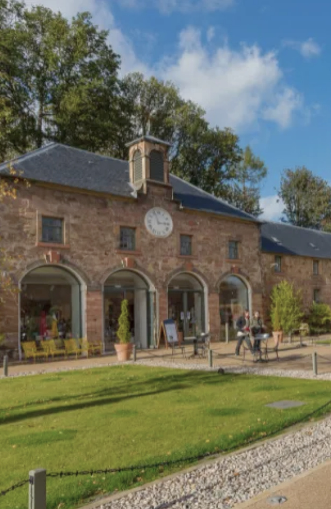 Historic brick building at the Restoration Yard in Dalkeith Country Park, with a clock above the arched entrances, outdoor seating, potted plants, and a gravel pathway surrounded by green grass and trees.
