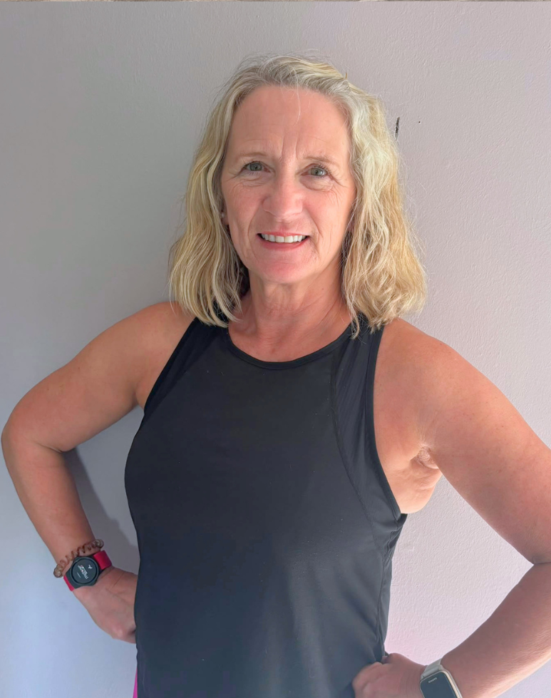 Instructor, Lynn, with shoulder-length blonde hair, smiling, wearing a black athletic tank top against a plain light-colored wall.