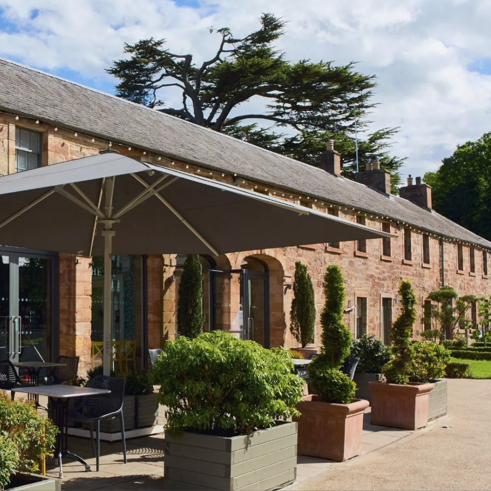 An outdoor patio area at the Restoration Yard in Dalkeith Country Park, with black chairs and tables, large potted plants, and a large umbrella in front of a stone building with multiple windows and trees in the background.