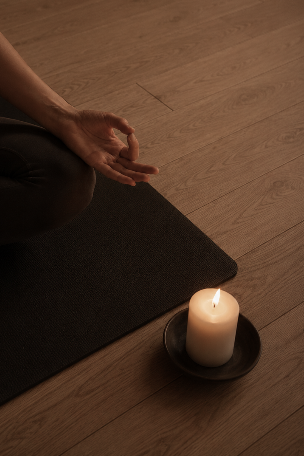 Person meditating sitting cross-legged on a yoga mat in dim lighting, with a lit candle on the floor nearby.