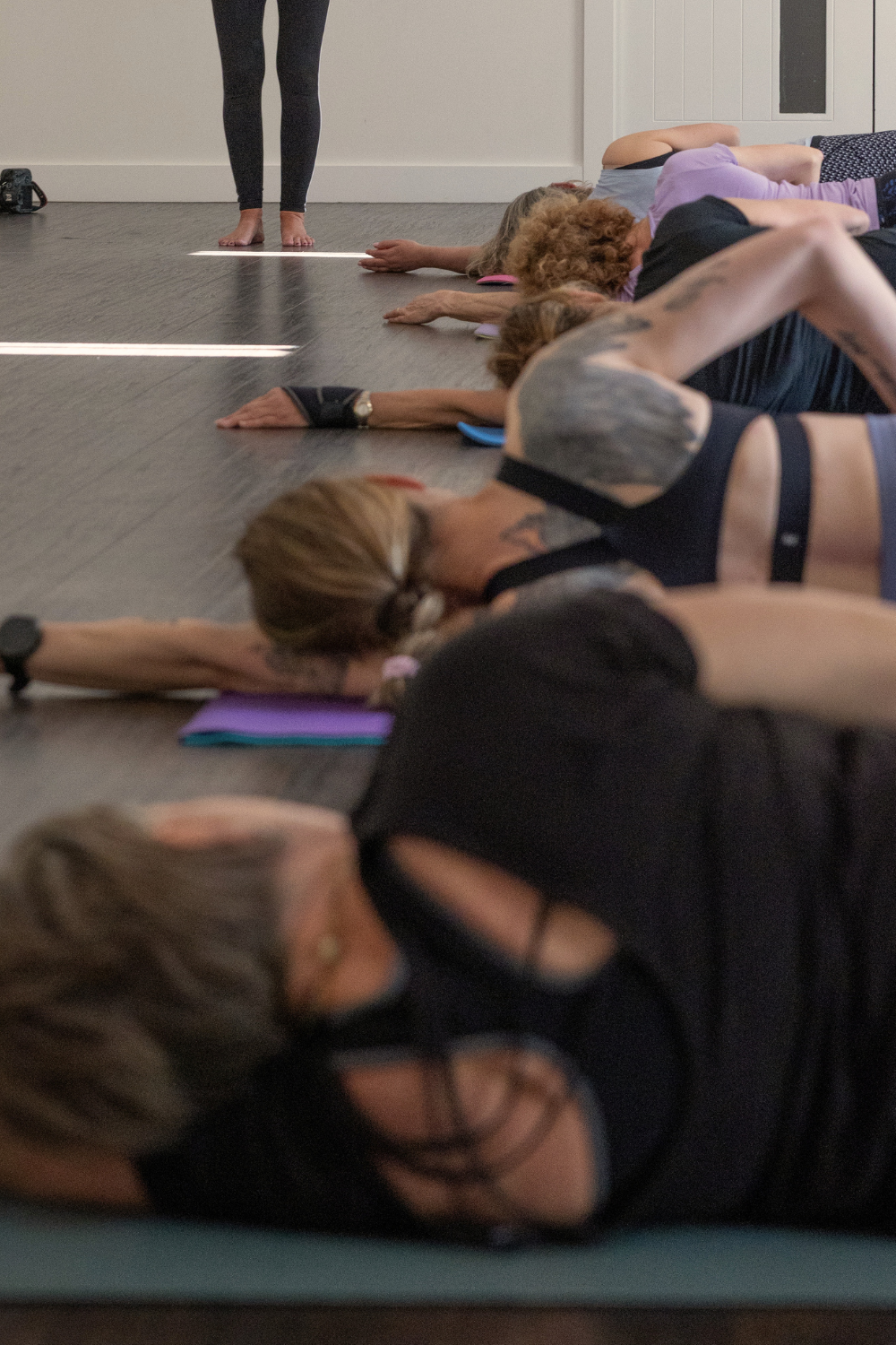 A group of women lying on the floor in a Pilates class, stretching with arms extended and heads resting on yoga blocks, with an instructor standing in the background.