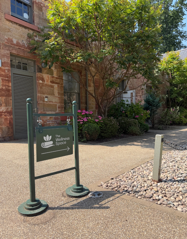 Signboard indicating the direction to The Wellness Space outside a brick building with trees and plants.