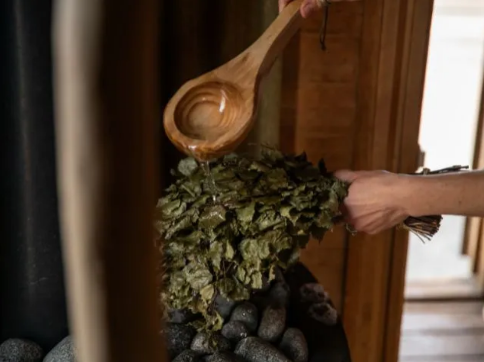 A person holding dried foliage with one hand while pouring water over it with a wooden spoon in a sauna setting.