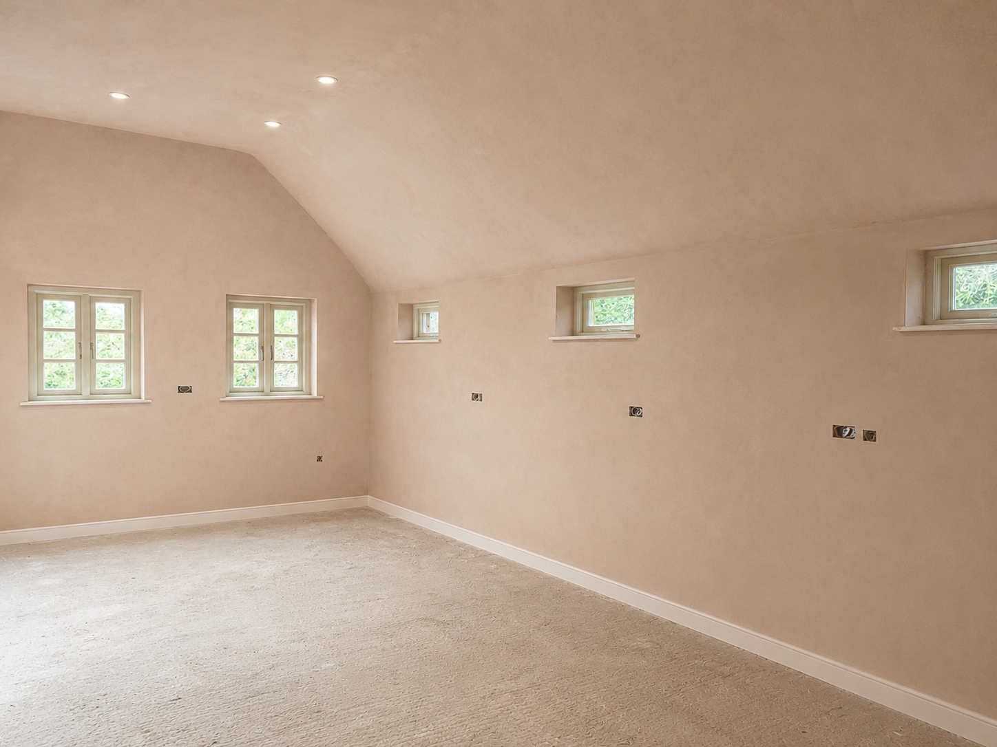 An empty room with beige walls, a vaulted ceiling, four small rectangular windows, and beige carpet flooring.