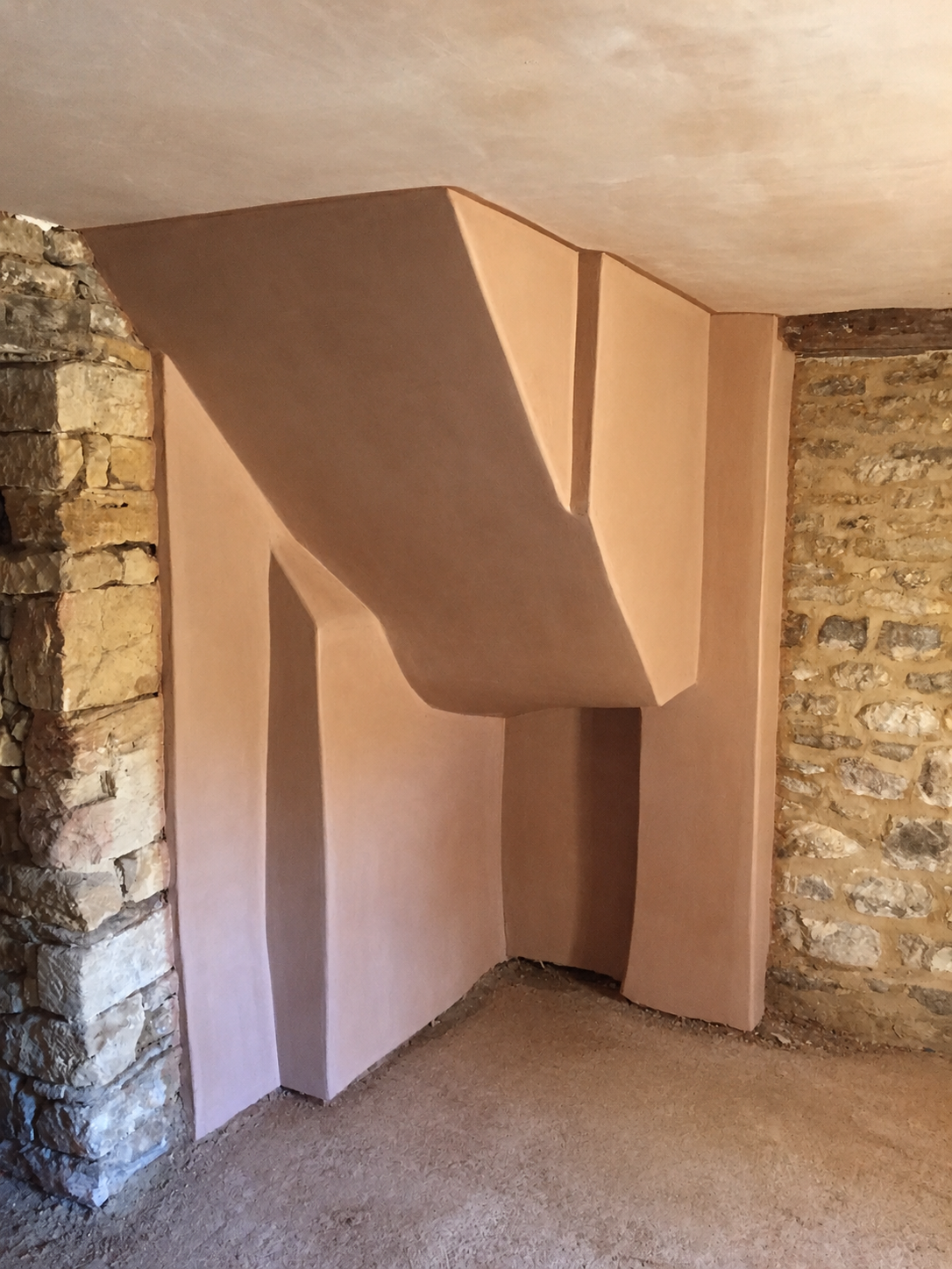 Interior view of a room with an unfinished curved wood structure or panel, showing rustic stone walls on the sides and a carpeted floor.