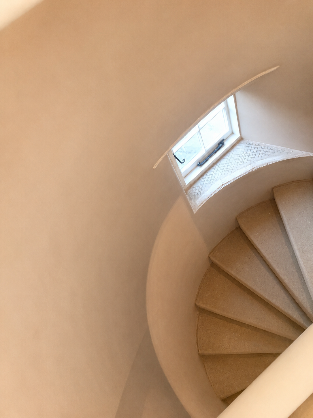 Spiral staircase seen from above with beige carpeted steps and a small window letting in natural light.