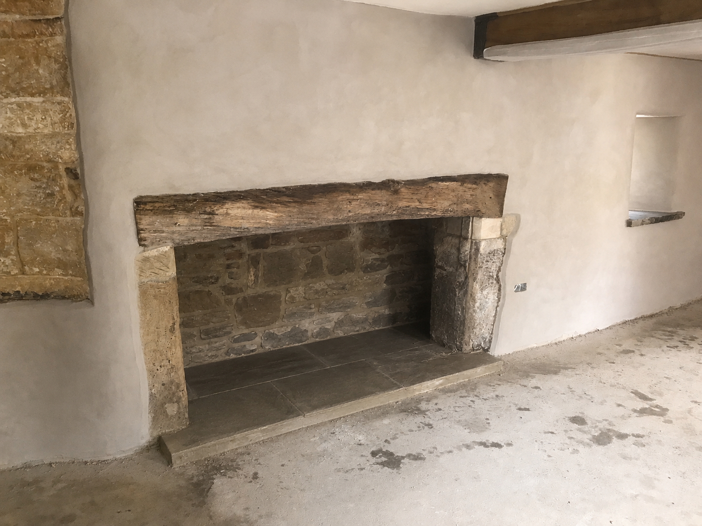 Empty fireplace with stone interior and rustic wooden mantel, in a room with plastered wall and small wall niches.