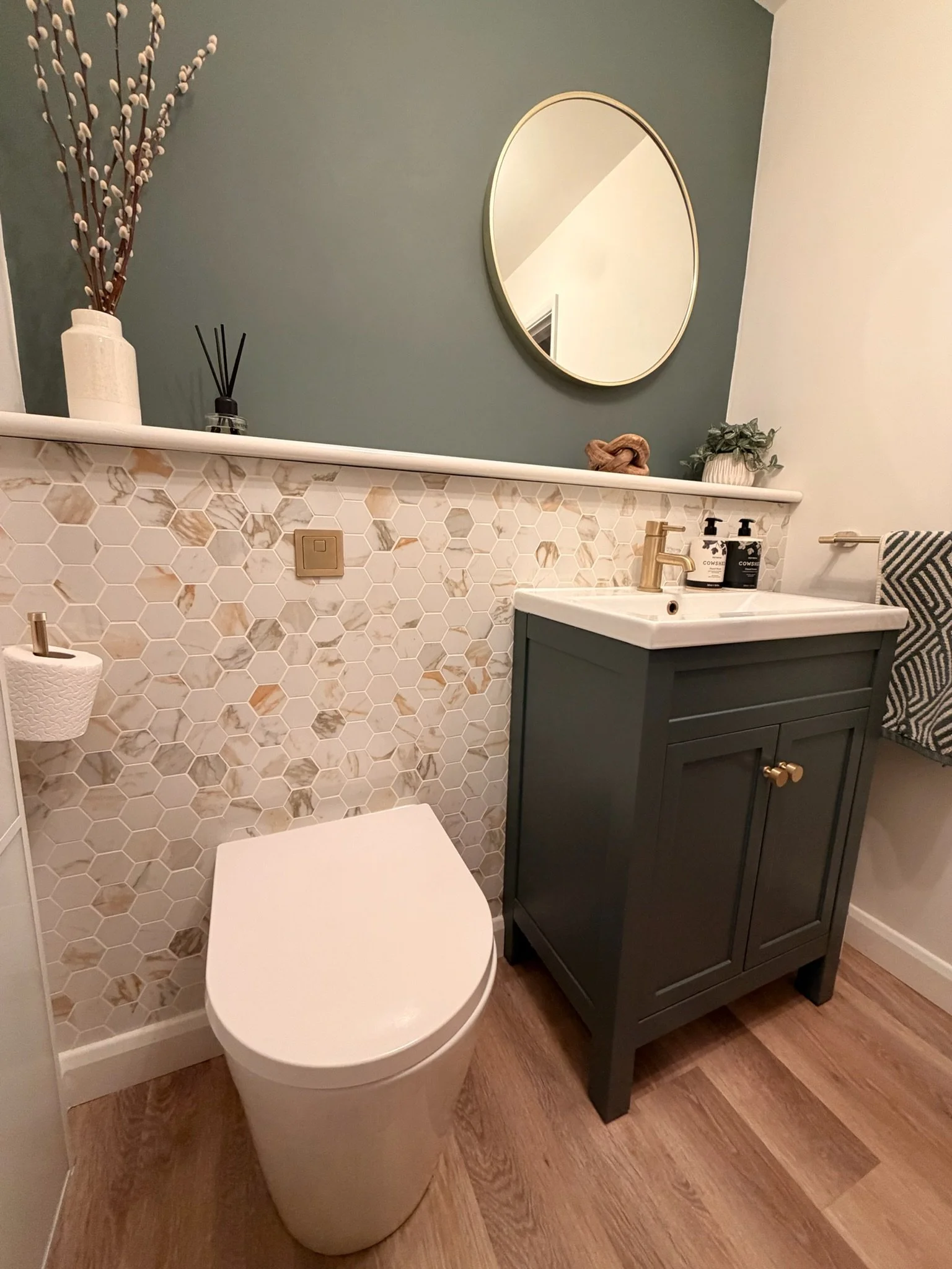A modern bathroom with a toilet, a green vanity with a white sink, a round mirror, decorative vases, and a wall with hexagonal marble tiles.
