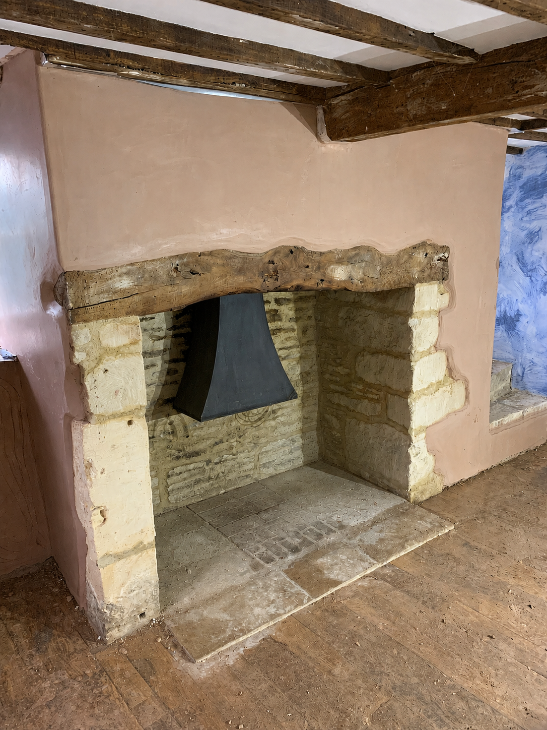 A rustic fireplace with a wooden mantel, stone brick surround, and a black hood, set against a pink plaster wall in a cozy room.