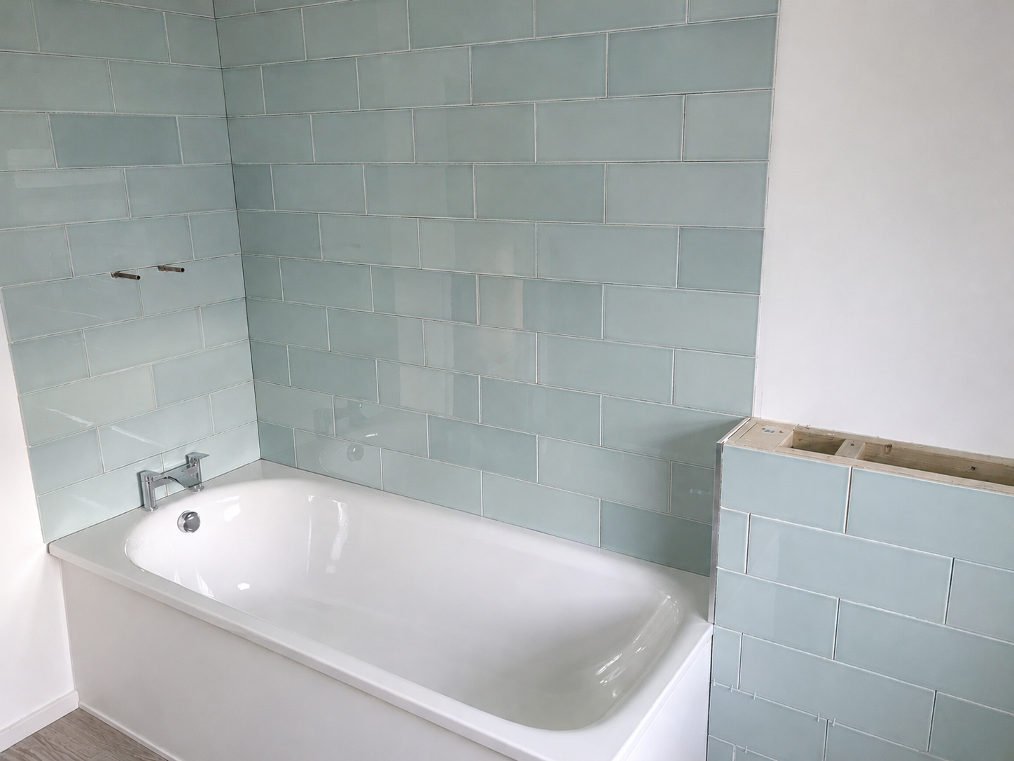 Bathroom with a white bathtub, light green subway tiles on the wall, and an unfinished section of wall or fixture on the right.
