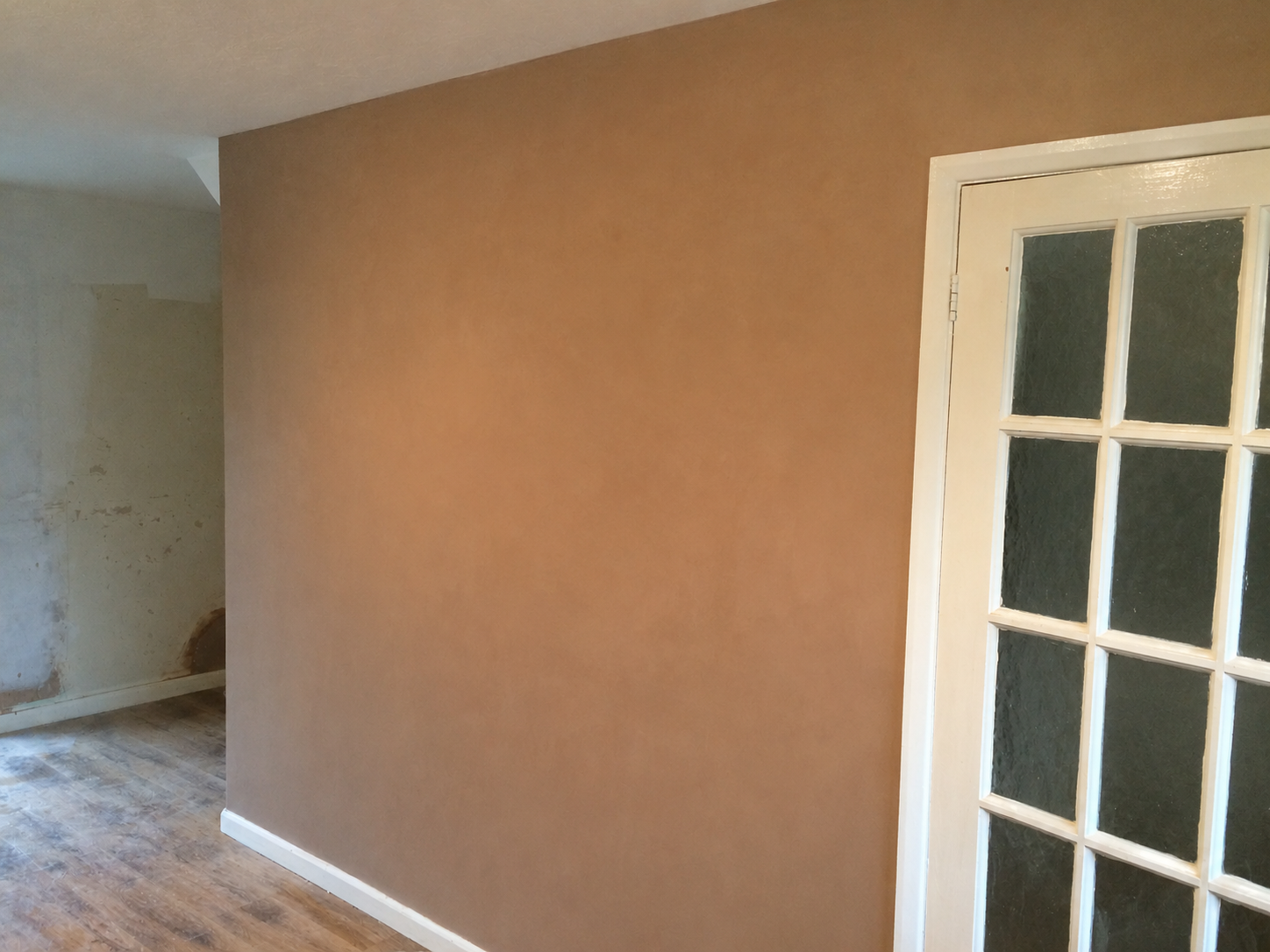 Interior room with a tan wall, a partially visible white-framed glass-paneled door, and a floor with wood pattern laminate. Another wall in the background shows a section with peeling paint and damage.