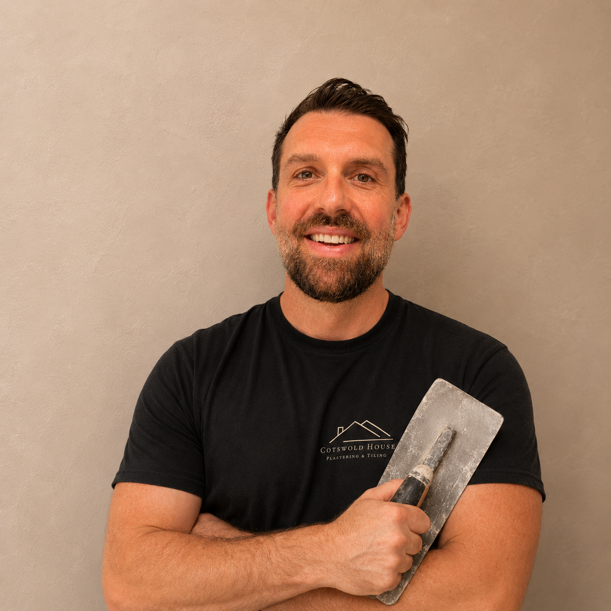 A smiling man with a beard holding a trowel against a plain beige wall
