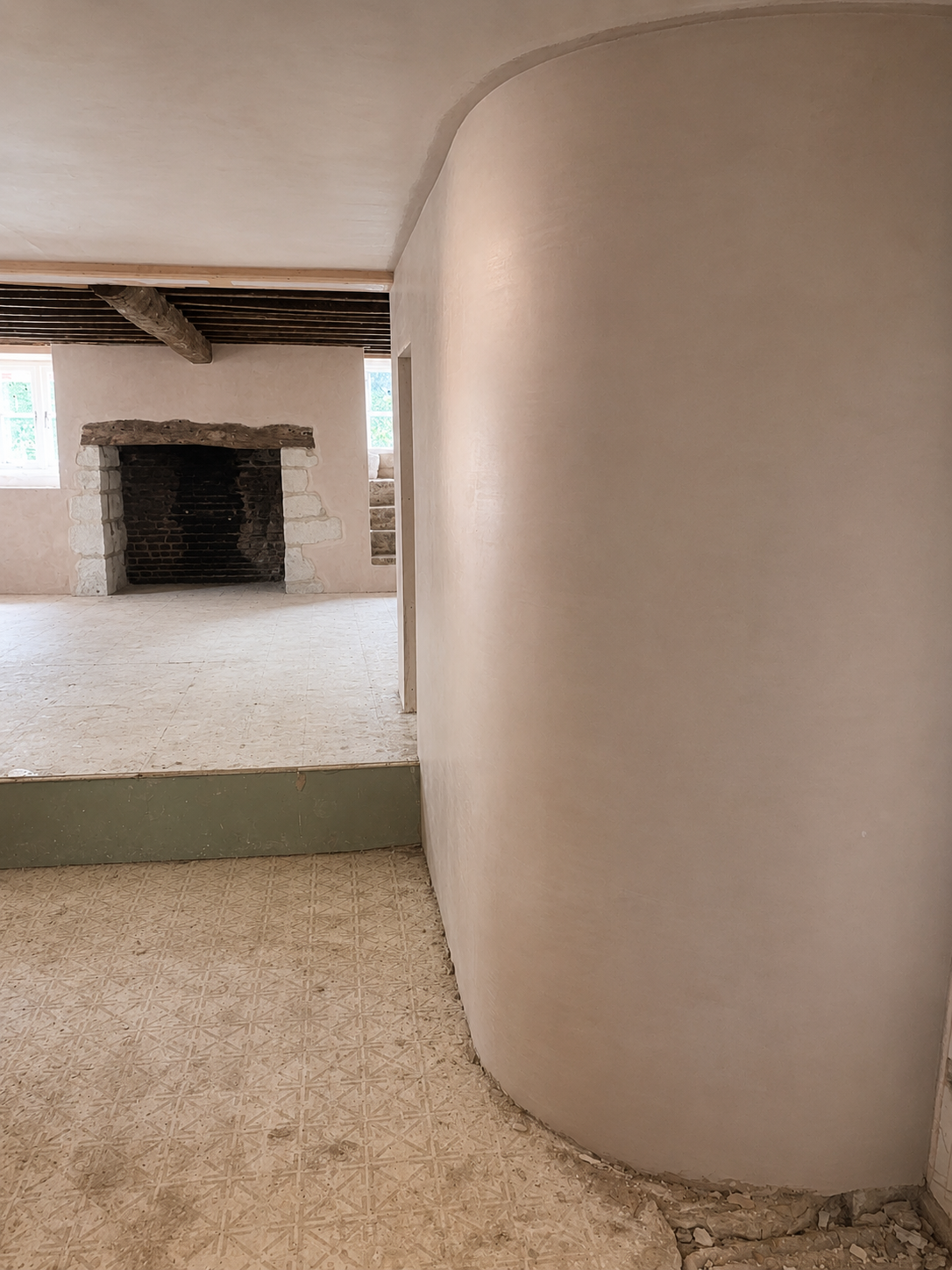 Interior of a room under renovation with a curved white plaster wall in the foreground, a stone fireplace in the background, visible window to the left, and construction debris on the floor.
