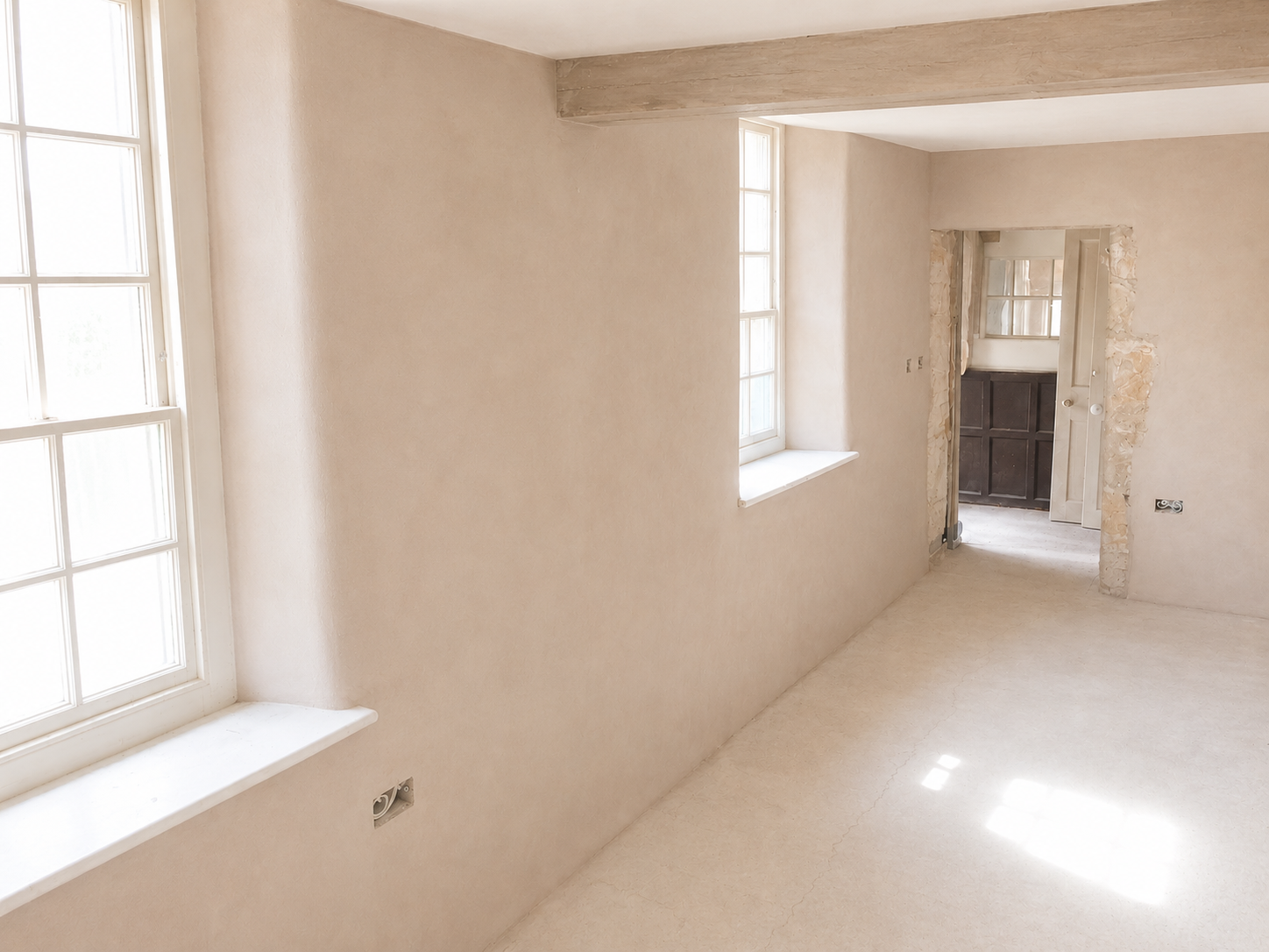 An empty room with light-colored walls, two large windows, and an unfinished doorway showing exposed brick and electrical outlets.