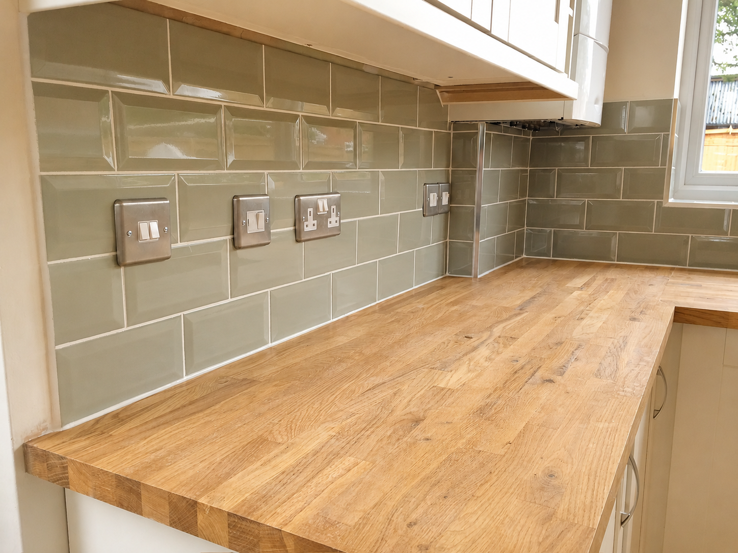 Kitchen countertop with olive green tile backsplash and multiple electrical outlets, next to a window.