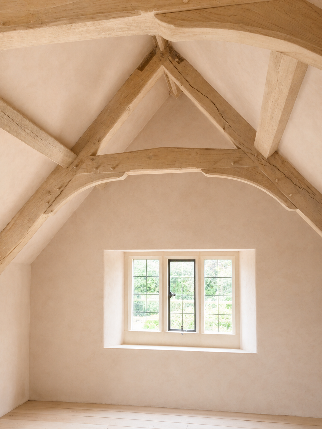 Interior view of a room with a high, vaulted ceiling featuring wooden beams, a large window with a tiny window on the side, and cream-colored walls.