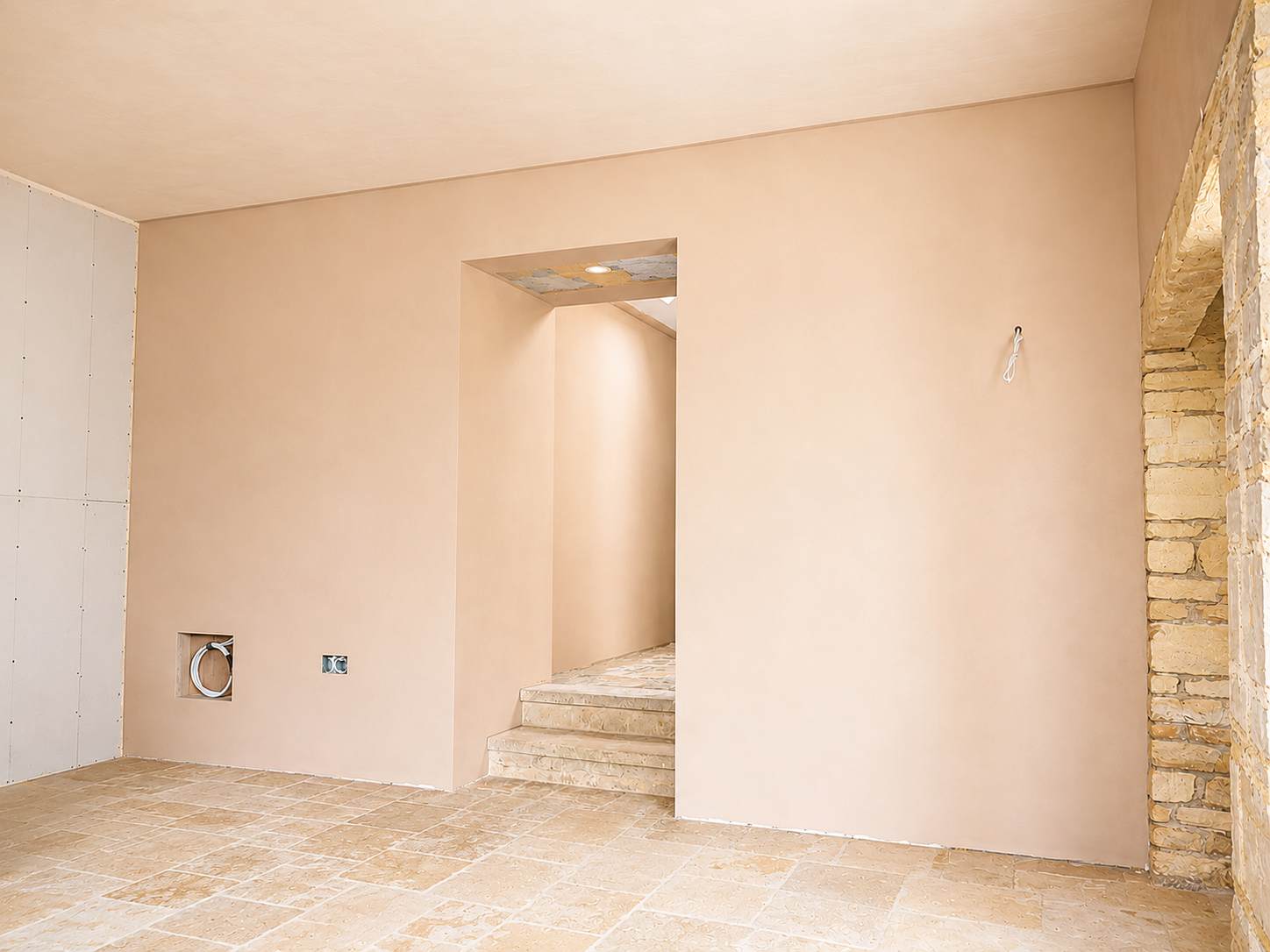 Empty room under construction with beige walls, unfinished ceiling, brick archway on the right, and plumbing outlets on the left wall.