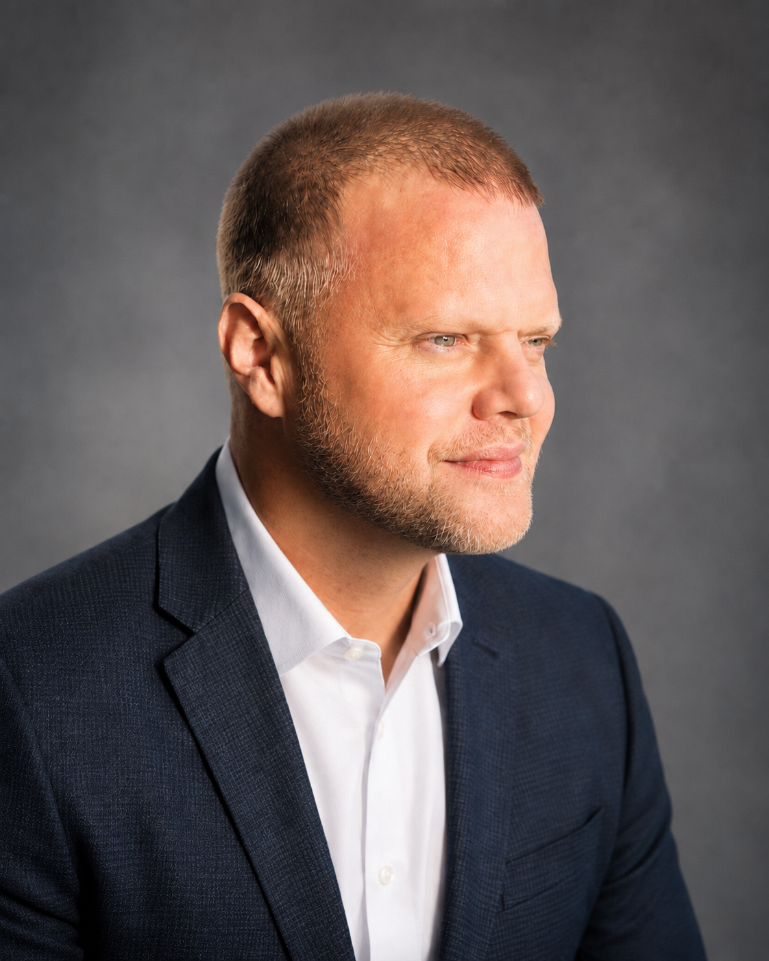 A portrait of a man with light skin, short light brown hair, and a beard, wearing a dark suit jacket and white shirt, against a gray background.