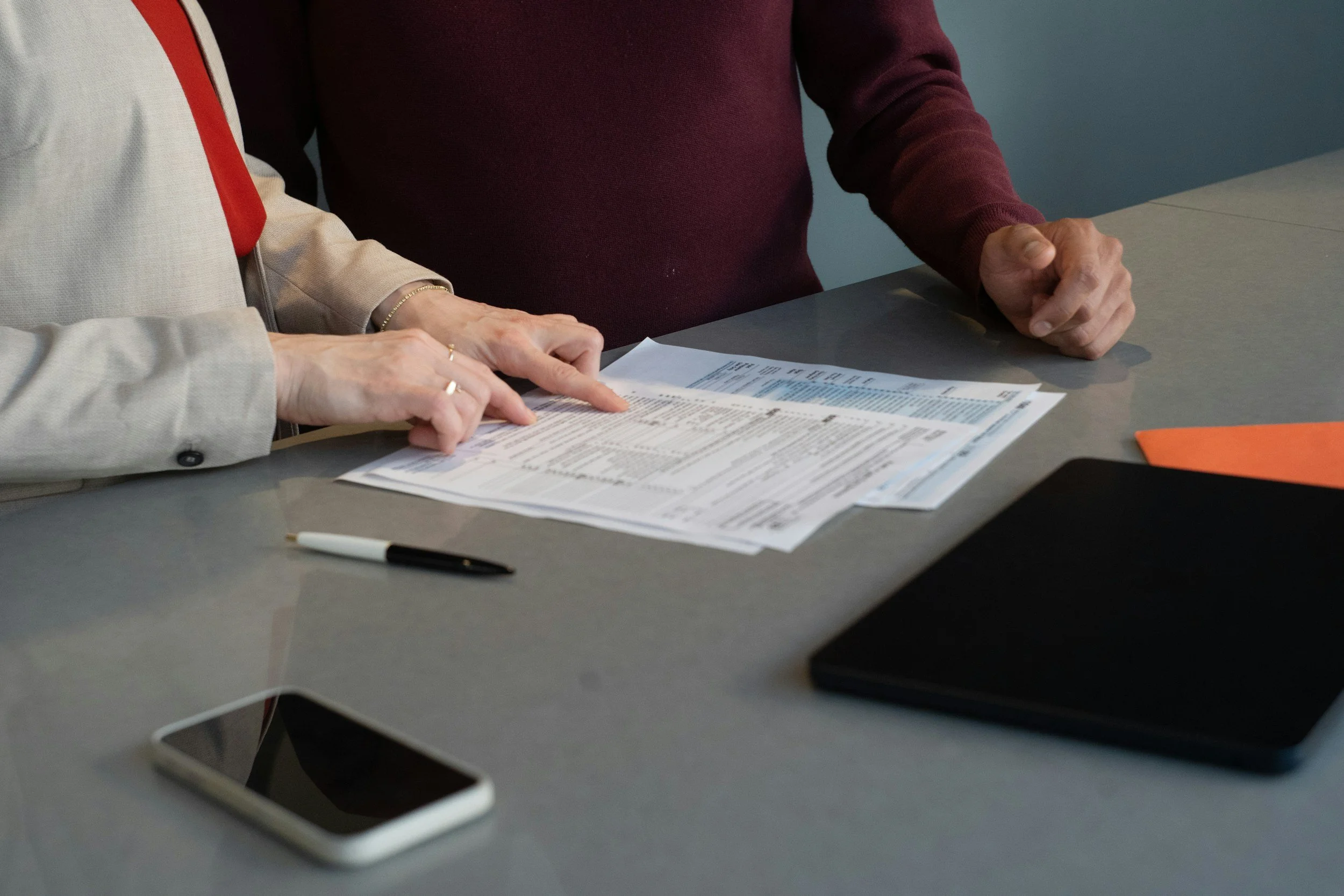 Two people are sitting at a table looking at a sheet with printed information. One person is wearing a light-colored blazer, while the other is in a dark red or maroon long-sleeve shirt. A pen and a smartphone are on the table.
