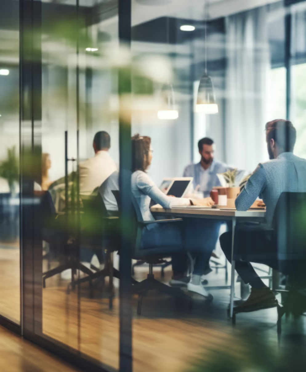 Business meeting through glass wall, people seated around table with laptops and documents, office setting.