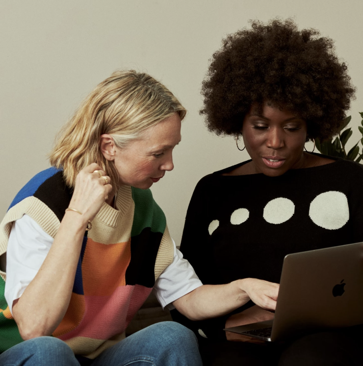 Two women sitting close together, looking at a laptop screen. One woman is blonde with a colorful sweater, and the other has dark curly hair with a black and white top.