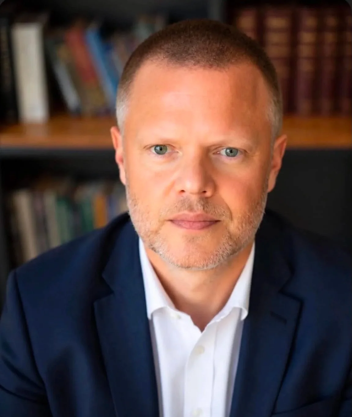 A middle-aged man with short brown hair and blue eyes wearing a navy blazer and white shirt, sitting in front of a bookshelf filled with books.