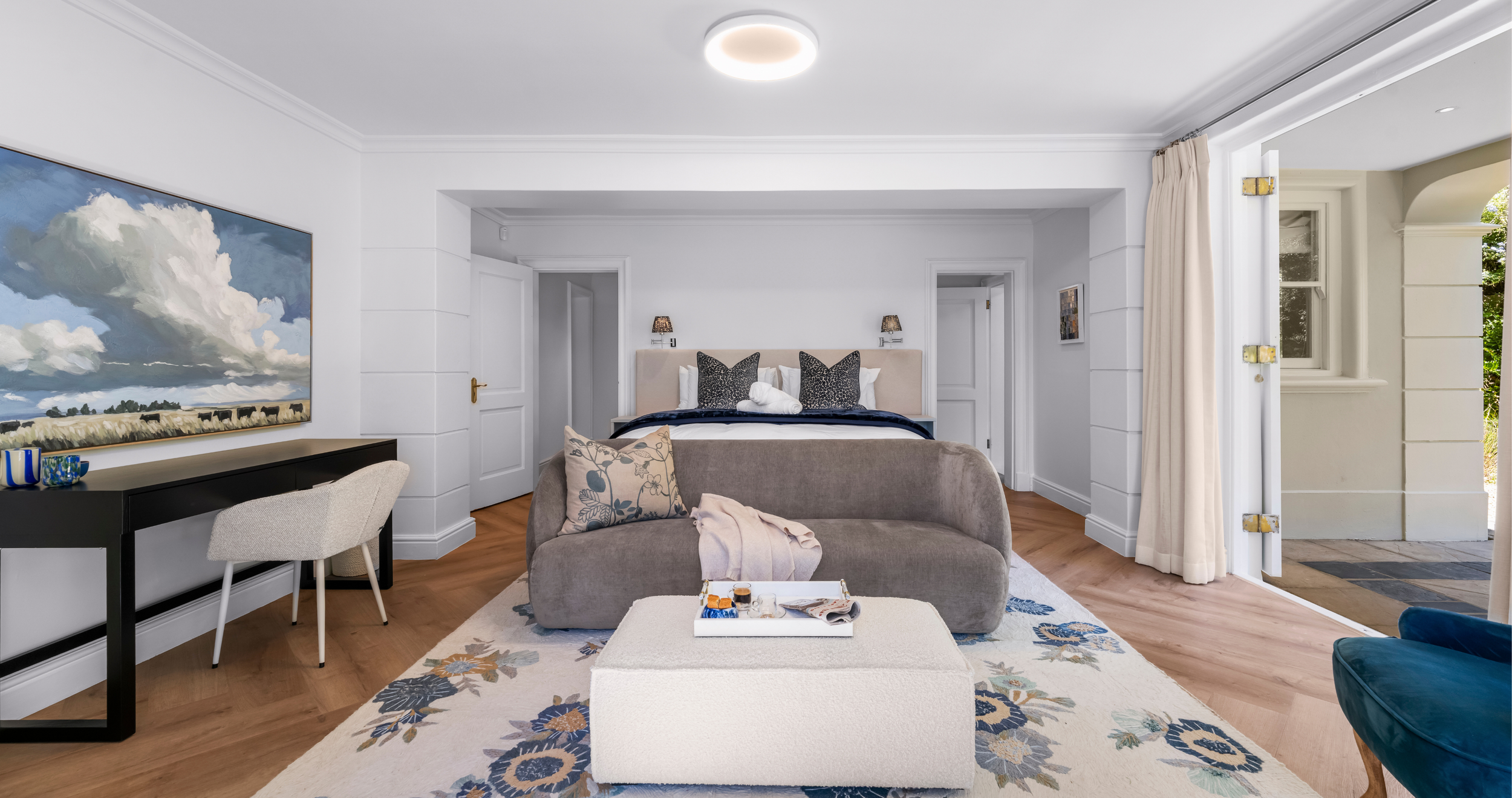Bright hotel bedroom with a bed, beige headboard, black and white pillows, and a rolled towel. A velvet gray sofa with patterned pillows is in front of the bed. There is a patterned area rug on wooden floor. A black console table with blue vases and 