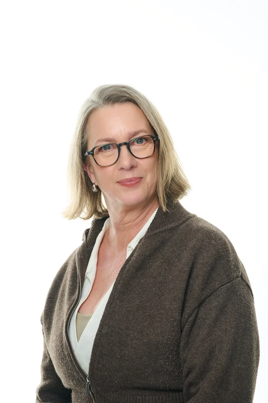 A middle-aged woman with shoulder-length gray hair, wearing glasses, a white blouse, and a brown zip-up sweater, with a neutral expression, against a white background.