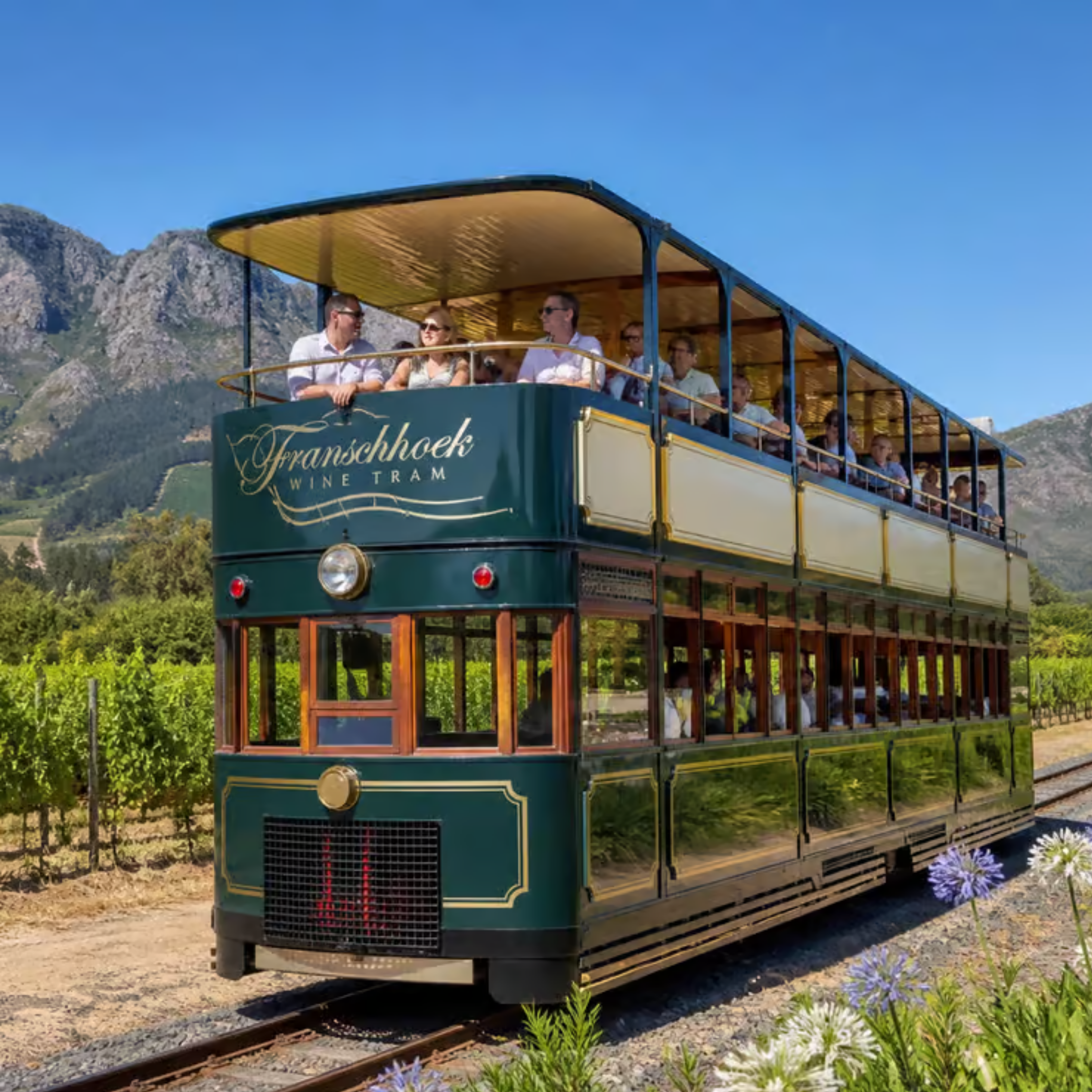 A vintage green two-story tram with open air upper deck carrying several passengers, traveling through a scenic mountainous landscape during daytime.