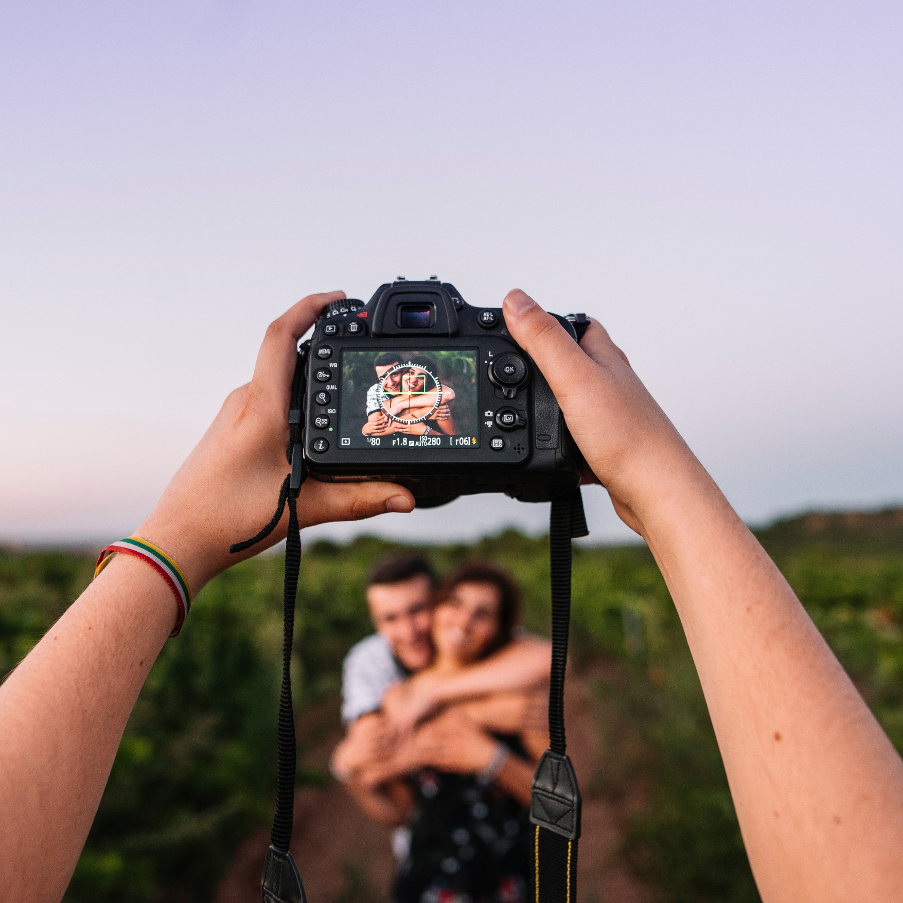 Photograph of a person taking a picture of a couple embracing, with the camera's viewfinder in focus showing the couple smiling and hugging, outdoors with a blurred green landscape background.