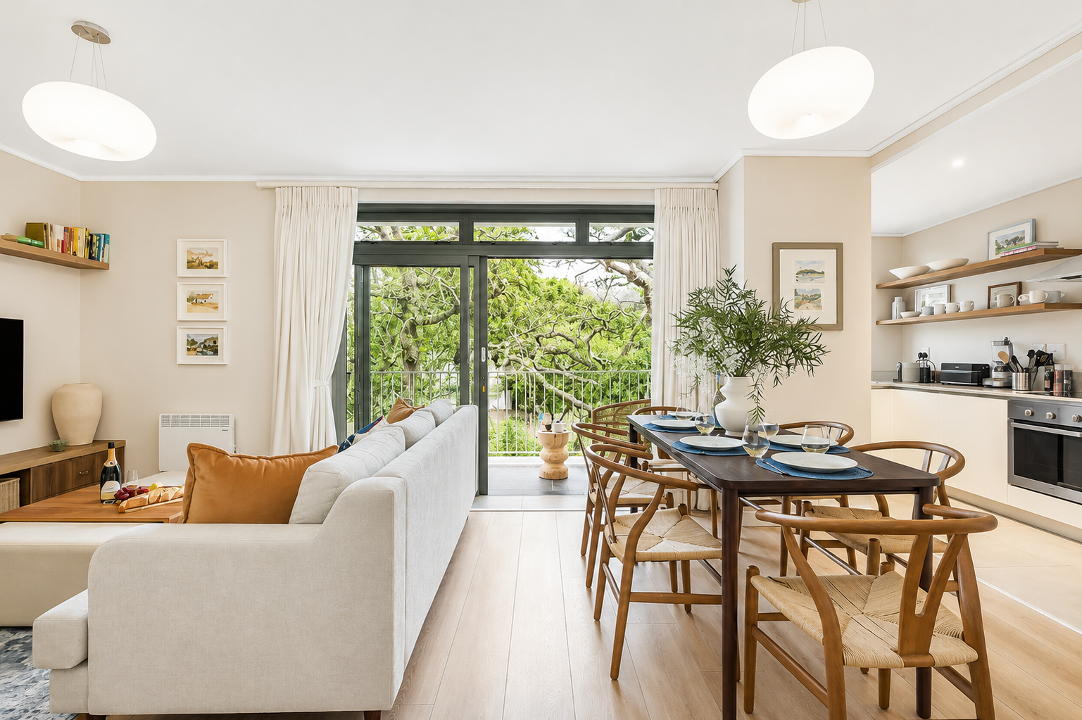 A living room and dining area with a large sliding glass door leading to an outdoor balcony surrounded by green trees. The interior has beige walls, a white sofa with tan pillows, a wooden dining table with six chairs, and open shelves with dishes in the kitchen.