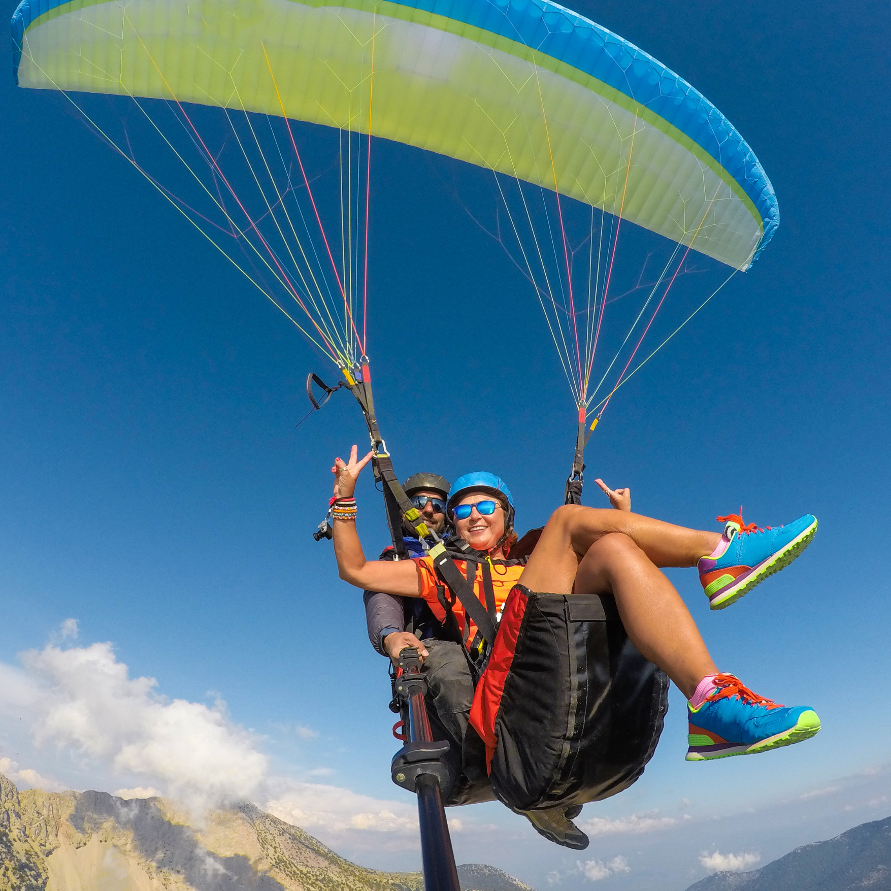 Two people paragliding high above a mountain landscape on a clear day with a blue sky and some clouds.