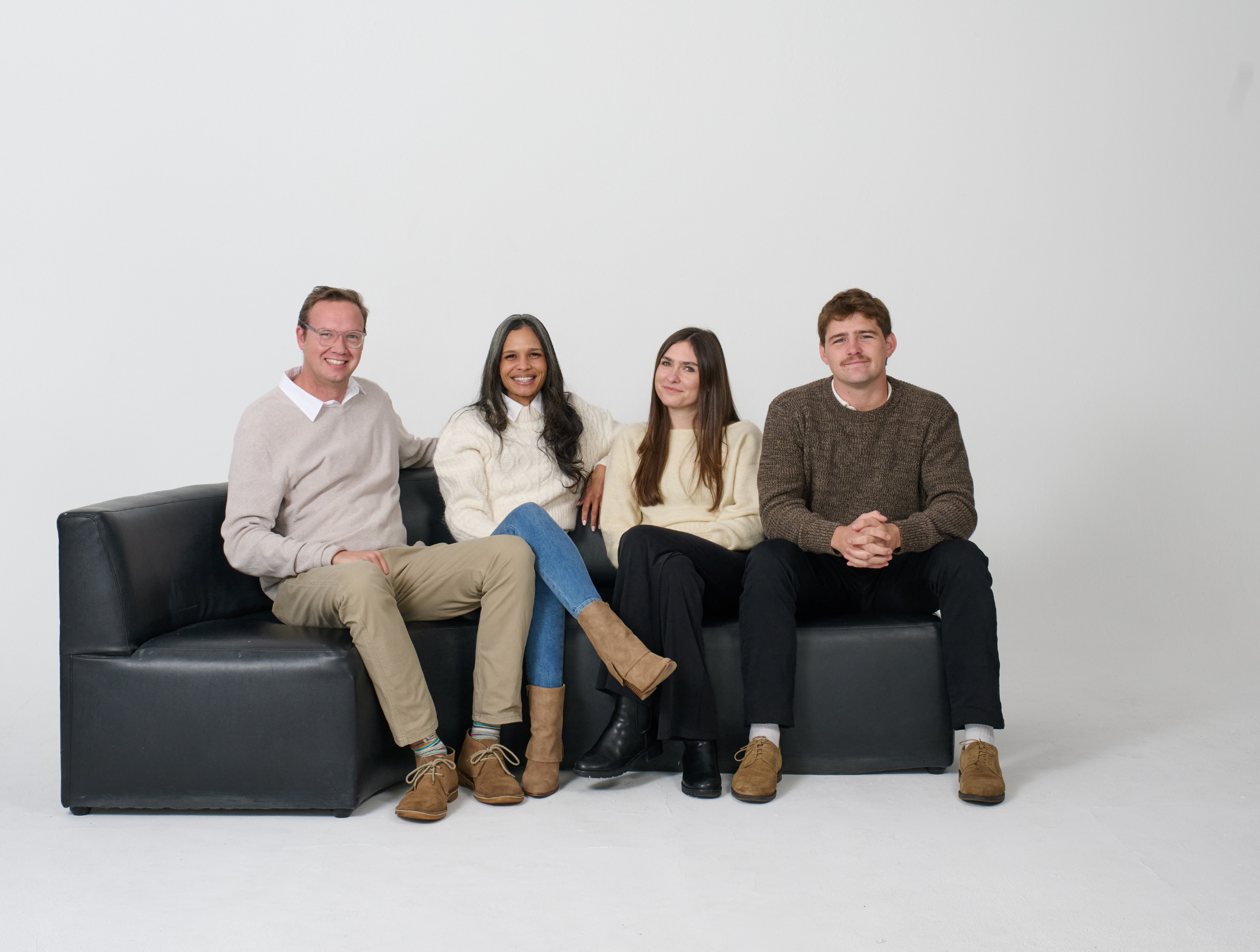 Four people sitting on a black leather couch against a plain white wall, dressed casually and smiling at the camera.