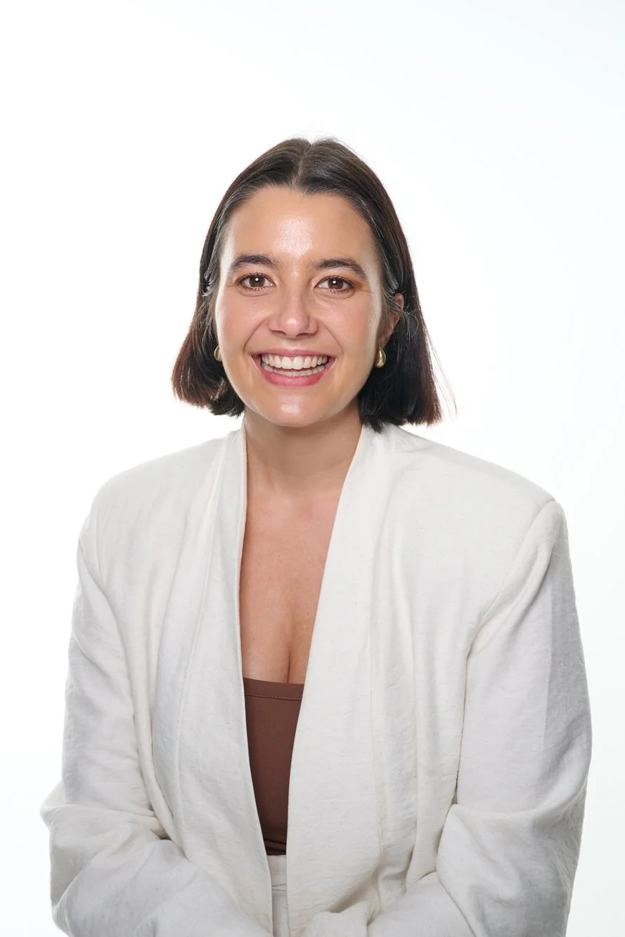 A woman with short dark hair, wearing a white blazer and a brown top, smiling against a plain white background.